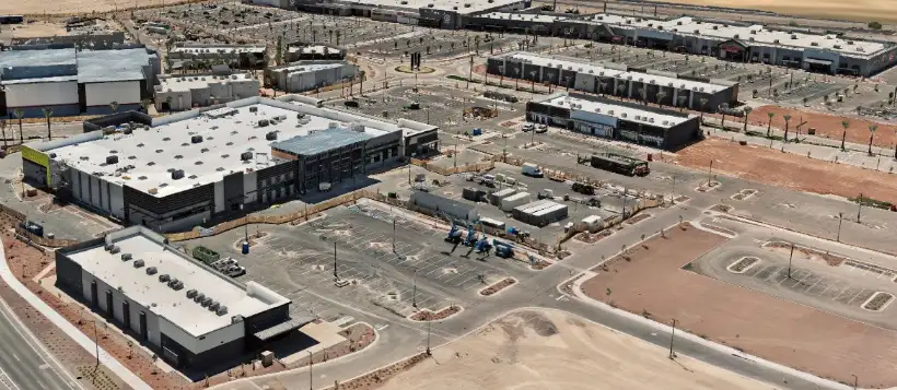 Oblique aerial photograph of Verrado Marketplace under construction, showing completed retail buildings with rooftop mechanical units, freshly striped parking areas, lifts and material staging on unfinished pads, and graded lots awaiting building.